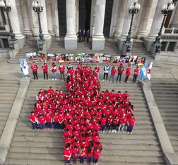 Argentine Red Cross volunteers wear red t-shirts and stand in the formation of a cross on the steps outside a public building. It is an aerial shot. 