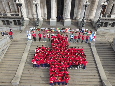 Argentine Red Cross volunteers wear red t-shirts and stand in the formation of a cross on the steps outside a public building. It is an aerial shot. 